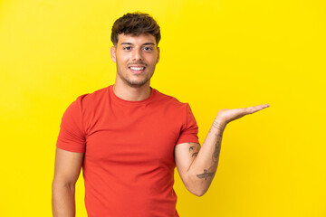 Young caucasian handsome man isolated on yellow background holding copyspace imaginary on the palm to insert an ad