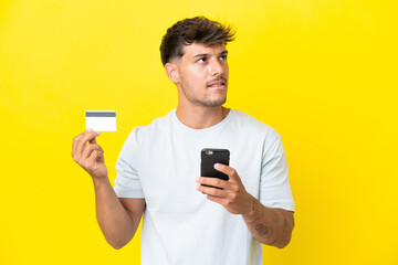 Young caucasian handsome man isolated on yellow background buying with the mobile with a credit card while thinking