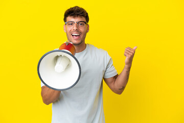 Young caucasian handsome man isolated on yellow background shouting through a megaphone and pointing side
