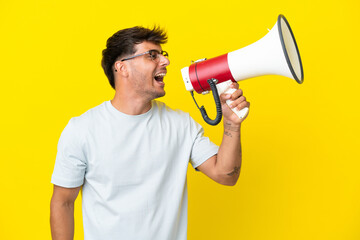 Young caucasian handsome man isolated on yellow background shouting through a megaphone to announce something in lateral position