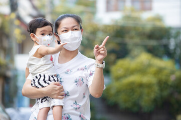 Obraz premium Asian mother wearing white face mask holding her little toddler baby girl who is her daughter wear baby face mask, pointing finger and looking out, COVID-19 concept