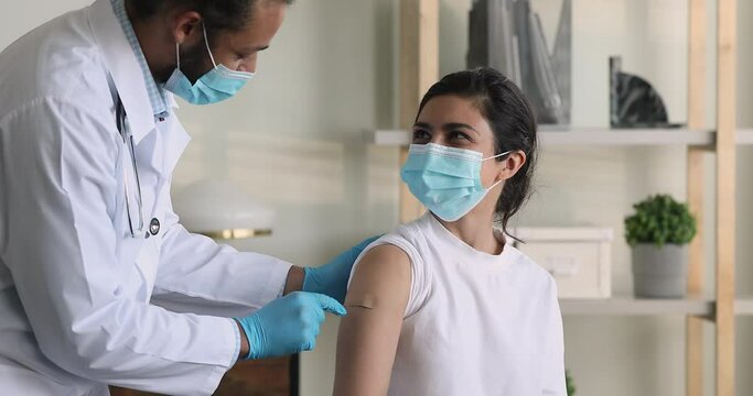 African male doctor wear surgical face mask protective gloves due precaution measures puts adhesive bandage on Indian female patient shoulder after injection against corona virus. Vaccination concept