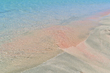 Blue sea water mixes white and pink sand on the beach.