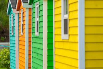 A row of small colorful painted huts or sheds made of wood. The exterior walls are colorful with double wooden doors. The sky is blue in the background and the storage units are sitting on gravel.  