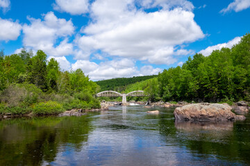 An old concrete archway bridge spans over a large salmon river with lots of rocks in the river.  There are tall green trees along both sides of the water. A hill or mountain is in the background. 