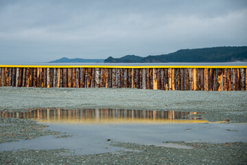 A breakwater made of wood logs and painted bright yellow on the top. There's an ocean and mountains over the wall. Before the seawater, there's a water puddle with the fence reflecting in the water.