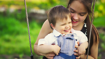 Cute brother and older sister ride on a bright swing on warm spring day against the backdrop of a green blooming garden