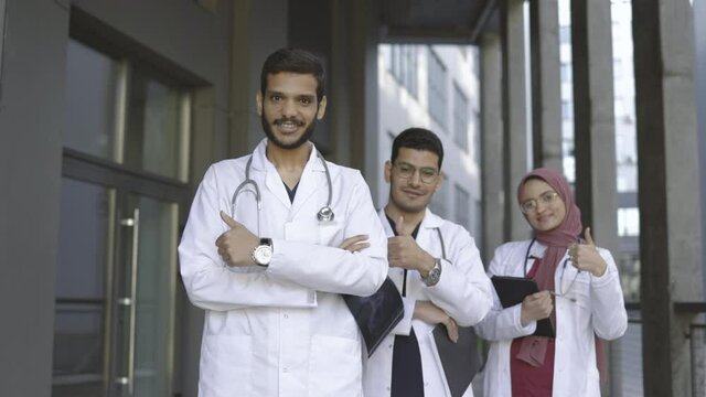 Healthcare and people concept. Happy successful Arab medical team in white lab coats standing with folded arms in a receding row and showing thumbs up, with focus handsome male doctor in front.