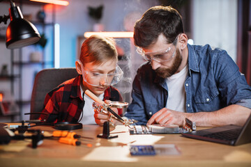 Happy little boy and his father sitting together at table and repairing display card using soldering iron. Two caucasian men wearing casual clothes and protective glasses.