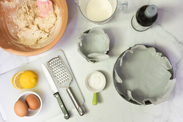 Baking ingredients for cheesecake on white table with copy space; top view. Eggs, flour, sugar, lemon, cream, creamcheese and kitchen utensils on table.