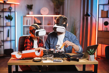 Young father helping his little son to fix remote controlled toy car during free time at home. Caucasian man and boy wearing modern VR glasses.