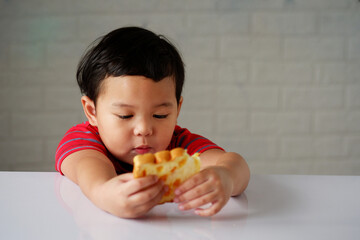 Cute little asian boy has an bored expression and is eating bread on a white dining table