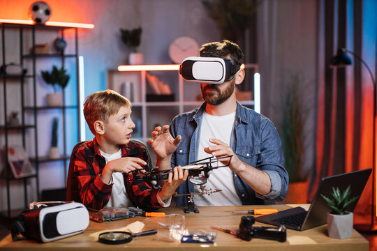 Little caucasian boy holding quadcopter while his father sitting near and wearing VR headset. Young father using modern gadget for fixing devices at home.