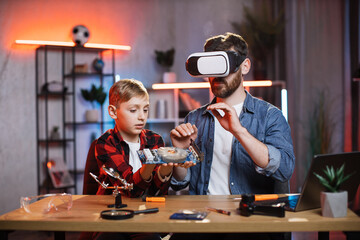 Young father in VR glasses helping his son to repair display card from modern laptop. Young man and little boy sitting at table and fixing details from computer.