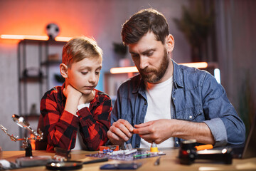 Caring caucasian father teaching his little son physics while assembling electric chain at home. Young man with boy using child designer toy for making experiment.