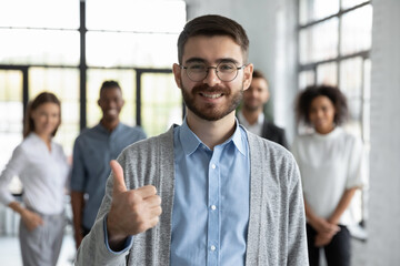 Portrait of smiling young Caucasian man in glasses stand in office show thumb up give recommendation. Happy male employee or client feel satisfied excited recommend good quality company service.