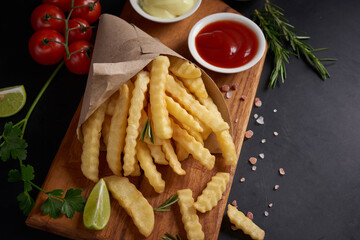 Homemade baked potato fries with mayonnaise, tomato sauce and rosemary on wooden board. tasty french fries on cutting board, in brown paper bag on black stone table background, unhealthy food.