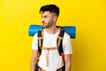 Young mountaineer caucasian man with a big backpack isolated on yellow background looking to the side