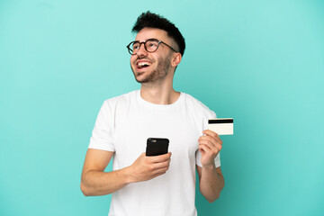 Young caucasian man isolated on blue background buying with the mobile with a credit card while thinking