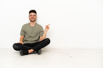 Young caucasian man sitting on the floor isolated on white background pointing finger to the side