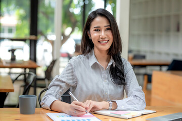 Charming young Asian businesswoman sitting at work holding pen using graph document at office. Looking at camera.