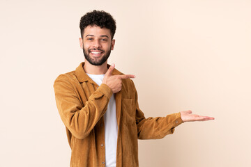 Young Moroccan man isolated on beige background holding copyspace imaginary on the palm to insert...