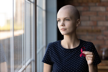 Thoughtful female oncology patient holding red ribbon, showing symbol of fight against breast...