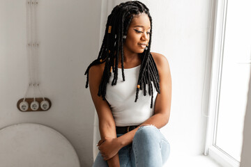 Gorgeous African American woman with cool dreadlocks in fashionable clothes sits near white wall and looks out window. Beautiful black afro girl with trendy hairstyle in fashion summer outfit indoors.