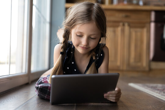 Smiling focused gen Z girl using app on tablet computer, resting on heating floor at home, watching movie or online learning event, reading book on internet. Childhood, distance education concept