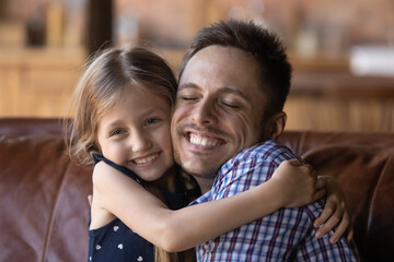 Happy daddy and cute little daughter girl hugging cheek to cheek with love and affection, sitting on couch at home, smiling. Kid embracing dad tightly, enjoying leisure time together. Family relations