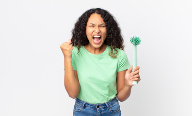 hispanic pretty woman shouting aggressively with an angry expression and holding a dish brush