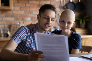 Happy young wife with cancer and husband doing routine domestic paperwork at home, paying bills,...
