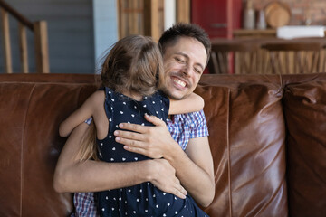 Happy young father hugging little preschooler daughter girl with love, care, affection, enjoying leisure time with kid at home, sitting on leather couch cuddling child. Best dad, family bonding