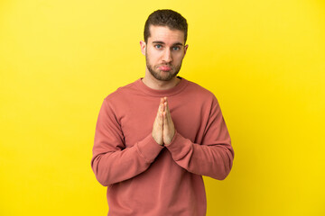 Handsome blonde man over isolated yellow background keeps palm together. Person asks for something