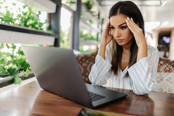 Young depressed woman sitting in cafe in front of laptop, looking at screen with frustrated expression, holding her head in hands, having headache, low or high arterial blood pressure, stress