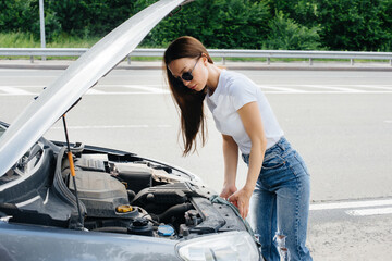 A young girl stands near a broken-down car in the middle of the highway and looks under the hood....