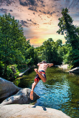 Unrecognizable Man Jumping From A Rock Into A Natural Water Pond Of A River With A Spectacular Sky. Young Man Enjoying A Summer Day In A Pond Of A River In The Middle Of Nature.He Jumps Into The Water