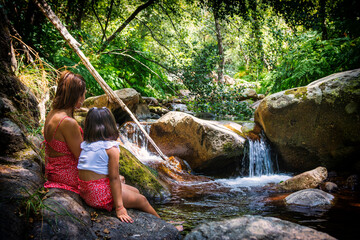 Mother And Daughter In Contact With Nature, They Are Sitting On A Rock Next To A River That Has A...