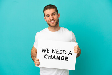 Handsome blonde man over isolated blue background holding a placard with text We Need a Change with happy expression