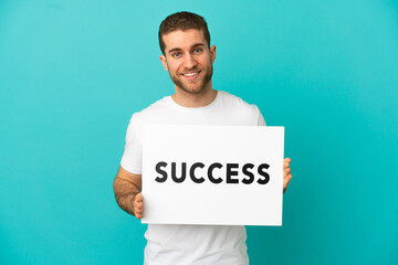 Handsome blonde man over isolated blue background holding a placard with text SUCCESS with happy expression