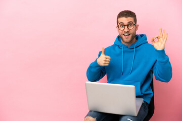Young man sitting on a chair with laptop showing ok sign and thumb up gesture
