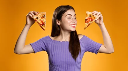Schilderijen op glas Happy smiling girl holding two slices of pizza, choosing what to eat, standing against orange background. Concept of pizzeria fast food, eating out and delivery © Liubov Levytska