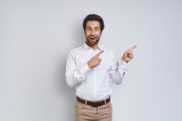 Handsome young smiling man in white shirt looking at camera and pointing away while standing against gray background