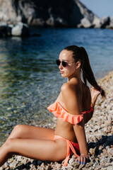 A beautiful young woman in glasses and a bikini is sitting on the ocean shore against the background of huge rocks on a sunny day. Tourism and tourist trips.