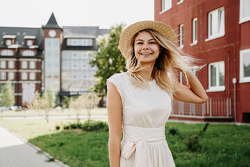 A beautiful blonde walks through a European city. Woman in white dress and straw hat, she smiles and looks at the camera