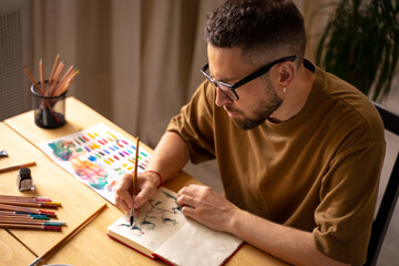 Portrait of Young dark haired concentrated caucasian white male artist with a beard in a glasses, khaki t-shirt. sitting in a studio in wooden table, holding a paint brush in his hand. DIY.