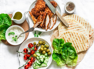 Ingredients for cooking greek gyros - chicken schnitzel, lettuce, grilled tortillas, greek yogurt tzatziki sauce, tomatoes, olives on a light background, top view
