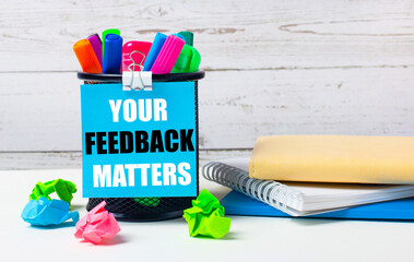 Against a light background of a wooden wall, there is a glass with colored markers, crumpled bright sheets of paper and a sheet of blue paper with the inscription YOUR FEEDBACK MATTERS