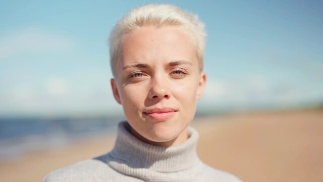 Slow motion portrait face of beautiful young woman with short blonde hair wearing gray turtleneck sweater and looking at camera standing on sea beach
