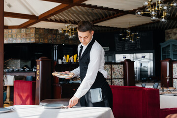 A young male waiter in a stylish uniform is engaged in serving the table in a beautiful gourmet restaurant. Restaurant activity, of the highest level.